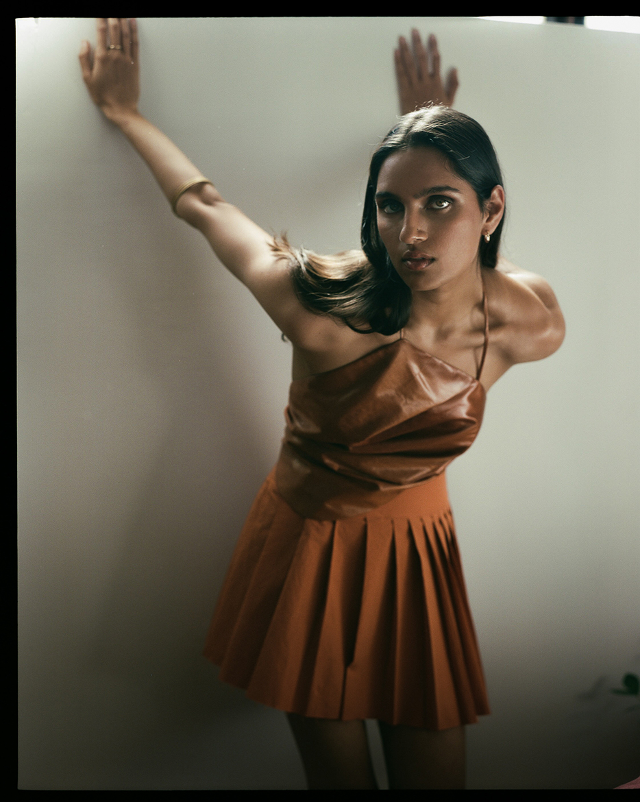  Model with their arms extended behind them leaning against a wall looking at the camera with smokey eye makeup on wearing a copper colored top and skirt agianst a neutral background